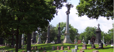 Image at St.John The Baptist Cemetery grounds headstones, trees, and flowers.