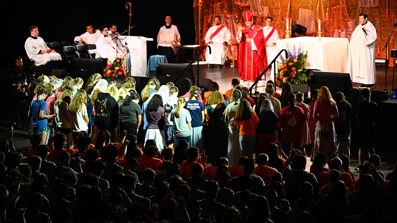 A group of young people standing in front of a lit stage and a man in red robes with his hands raised in an auditorium full of people